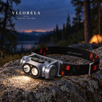 Gray dual-beam rechargeable headlamp at a forest campsite after dusk.